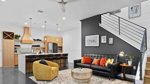 A modern living room with a sofa, a yellow armchair, and a coffee table. There is a kitchen area with wooden cabinets and a bar counter. A staircase with a black railing is visible in the background. - South Lamar Luxe Creekside Hot Tub (Austin)