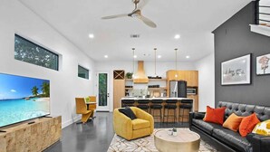 A modern living room with a large TV, a sofa, a coffee table, and a dining area with a bar. - South Lamar Luxe Creekside Hot Tub (Austin)