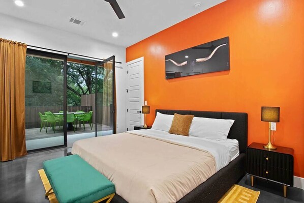 A modern bedroom with a large bed, a glass door leading to an outdoor seating area, and an orange accent wall. - South Lamar Luxe Creekside Hot Tub (Austin)