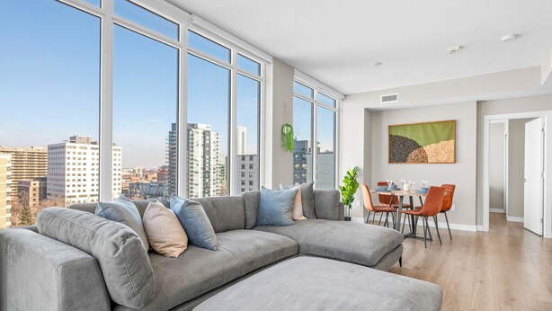 A modern living room with a large sectional sofa, a dining area with a table and chairs, and large windows offering a city view.