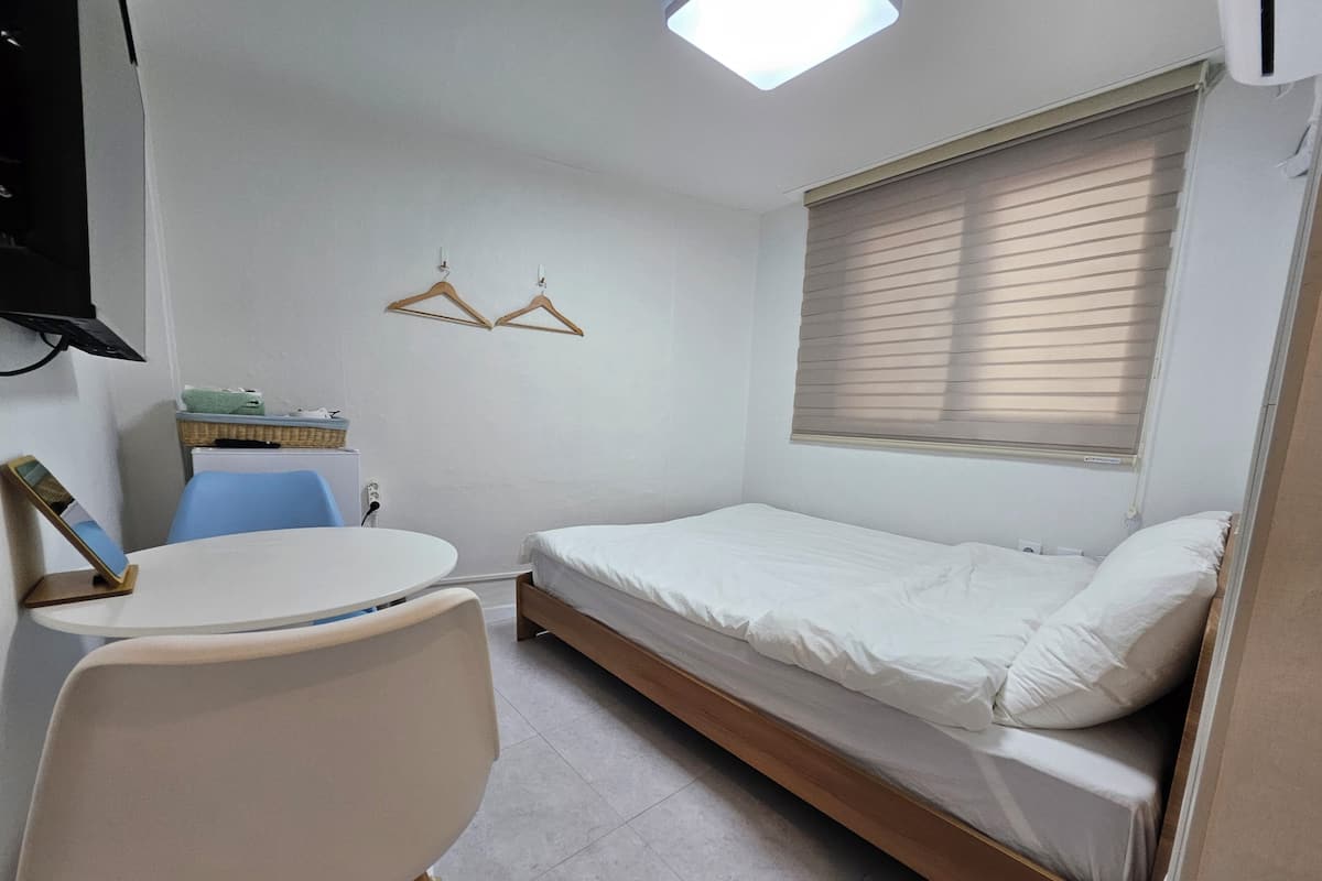 Habitación compacta con una cama individual, una mesita con una silla, un televisor montado en la pared y una ventana con persianas.