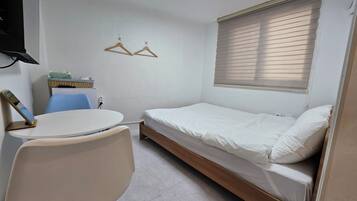 A compact room with a single bed, a small table with a chair, a TV mounted on the wall, and a window with blinds.