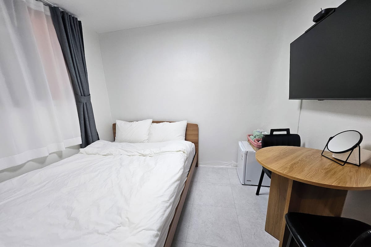 Una habitación de hotel con cama, escritorio con silla, refrigerador pequeño y televisor fijado a la pared.