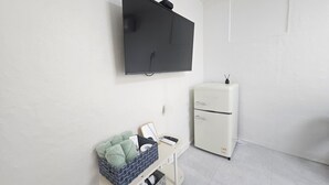 A room with a flat-screen TV mounted on the wall, a white refrigerator, a small table with a basket of green towels, and a white cabinet. - Daldal Masil (Mokpo)