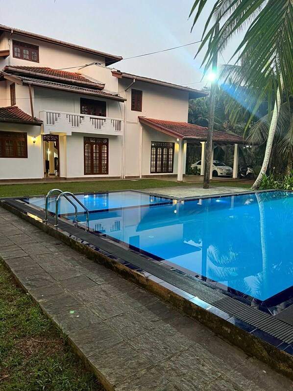 A swimming pool with a tiled deck, a covered seating area, and a building with a balcony in the background.
