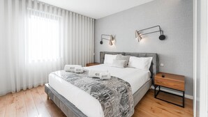 Quarto de hotel moderno com uma cama grande, paredes cinza texturizadas e duas mesas de cabeceira com luminárias.