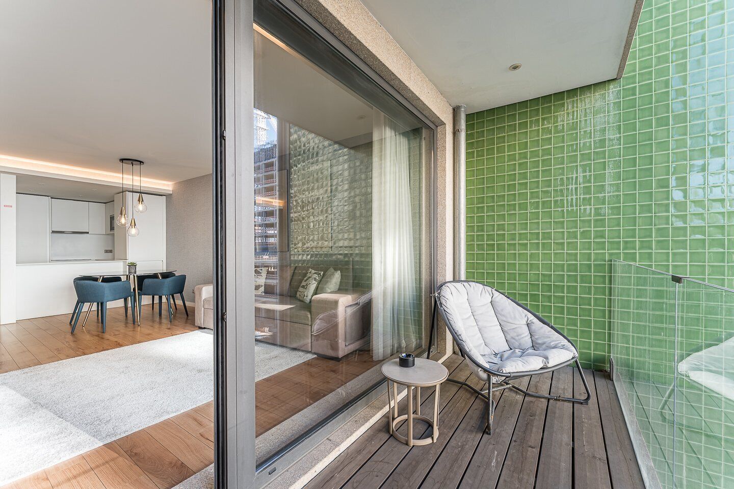 Un salon moderne doté d’une porte vitrée donnant sur un balcon, d’un coin repas avec des chaises bleues et d’un mur recouvert de carreaux verts.