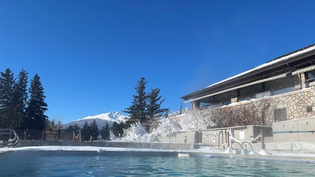 A pool with clear blue water, a stone building with a stone facade, and a snow-capped mountain in the background. - Hinton Mountain House (Hinton)