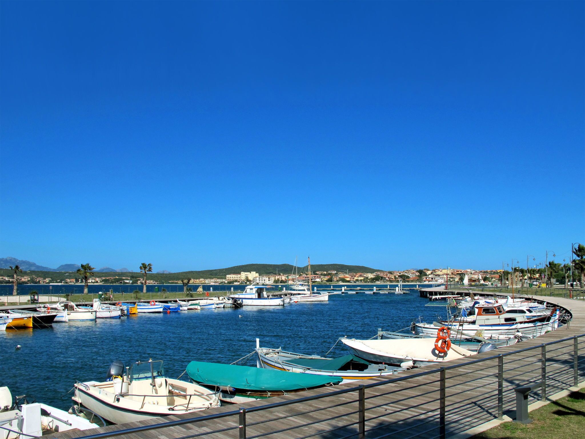 Een jachthaven met verschillende boten die liggen aangemeerd, een heldere blauwe lucht en een heuvelachtig landschap op de achtergrond.