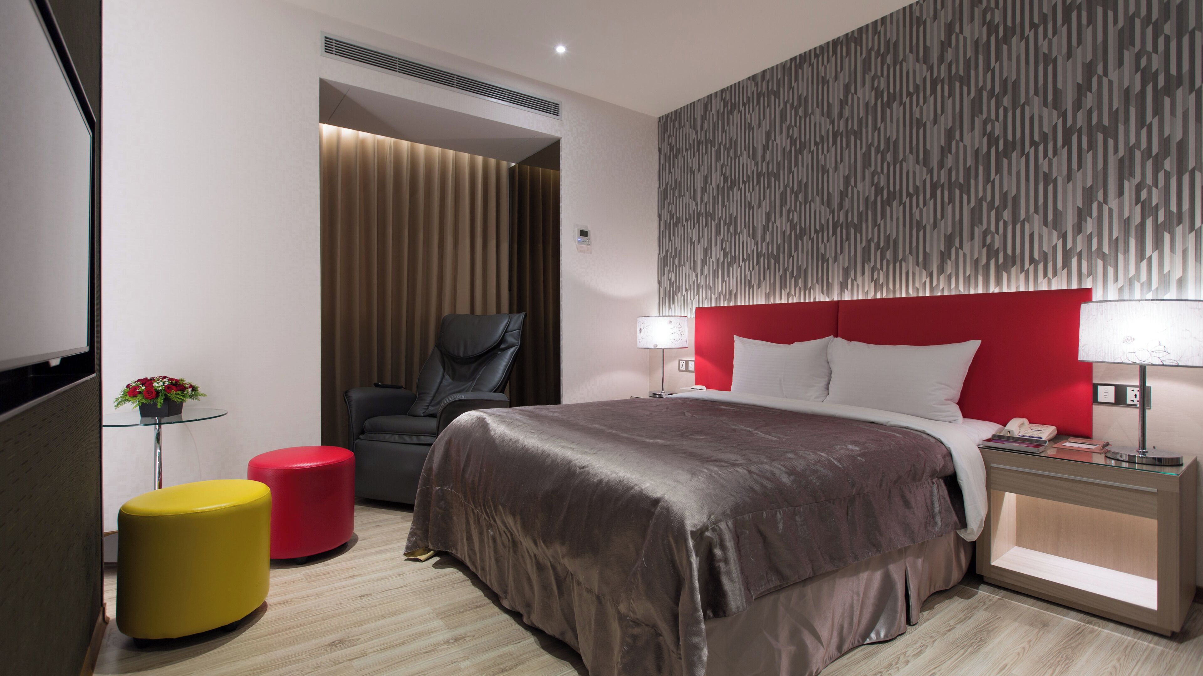 Une chambre d’hôtel moderne avec un grand lit, un mur d’accent rouge, une table en verre avec un tabouret rouge et un pouf rouge.