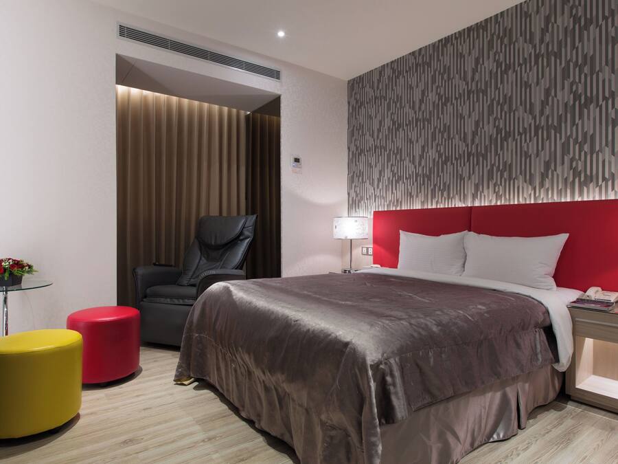 A modern hotel room with a large bed, a red accent wall, a glass table with a red stool, and a red ottoman.