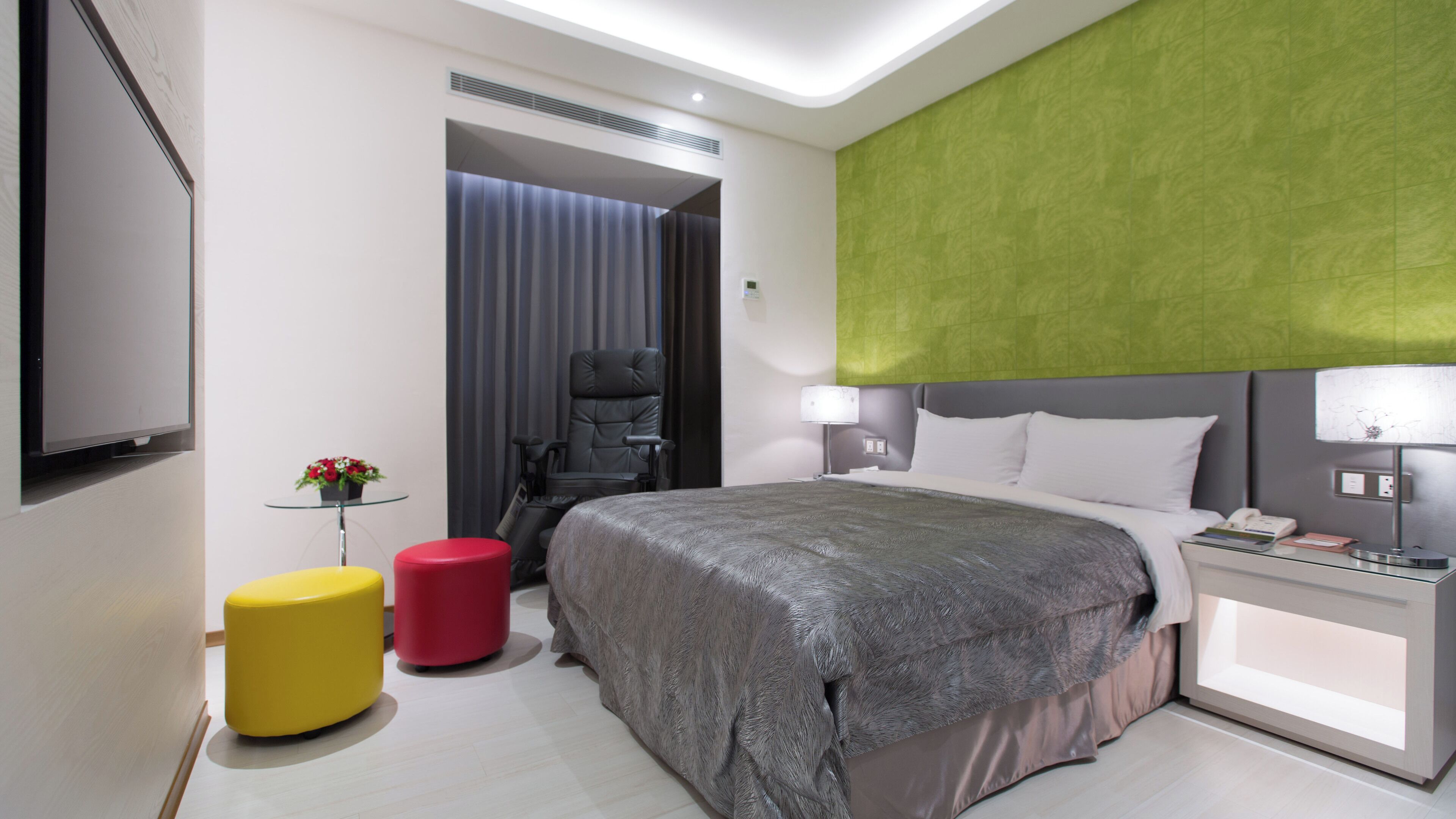 A modern hotel room with a large bed, a desk with a lamp, a chair, a small table with a plant, and a TV.