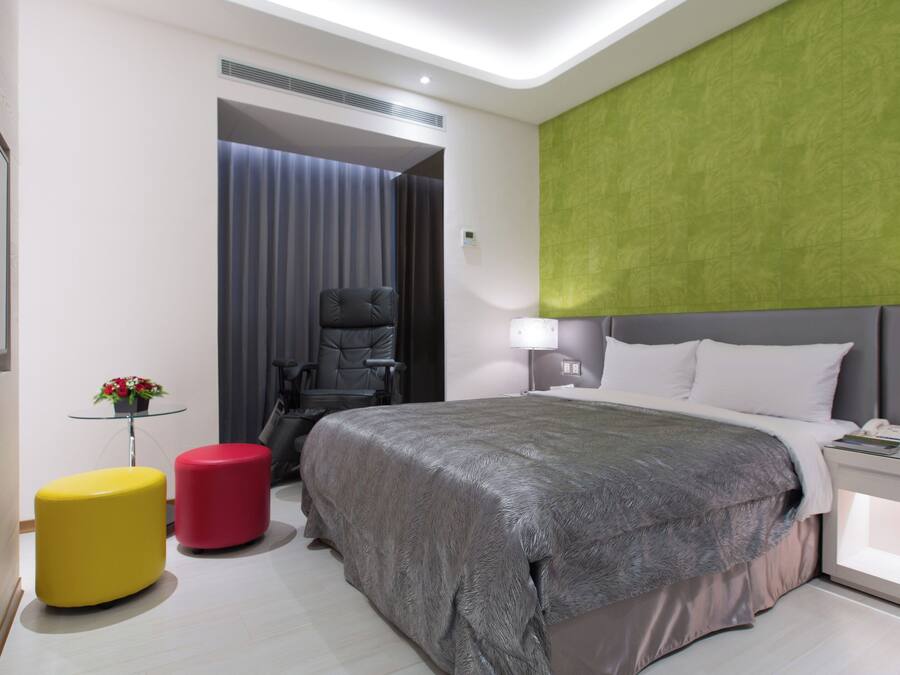 A modern hotel room with a large bed, a desk with a lamp, a chair, a small table with a plant, and a TV.
