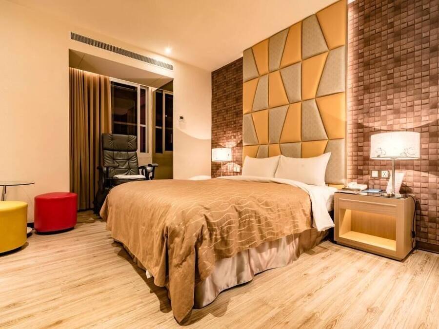 A hotel room with a large bed, a red ottoman, a nightstand with a lamp, and a brick-patterned wall.