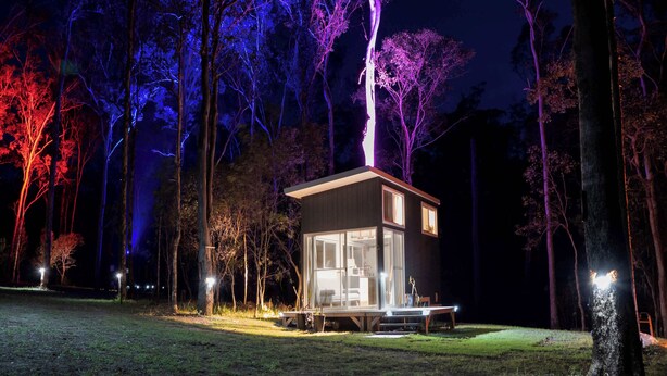 A modern, two-story cabin with a glass facade, surrounded by trees and illuminated by ambient lighting.