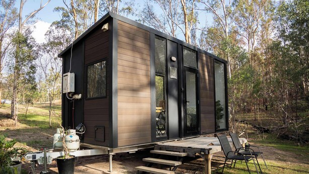 A modern, compact cabin with a deck and outdoor seating area, surrounded by trees.