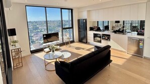 A modern living room with a black sofa, a coffee table, and a TV. There is a large window offering a city view. - The Luxury Penthouse at Spring Square (Bankstown)