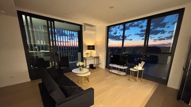 A modern living room with a black sofa, a round coffee table, and a TV. Large windows offer a city view. - The Luxury Penthouse at Spring Square (Bankstown)