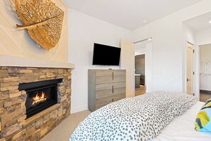A modern bedroom with a stone fireplace, a large bed with patterned bedding, a wooden dresser, and a flat-screen TV mounted on the wall.
