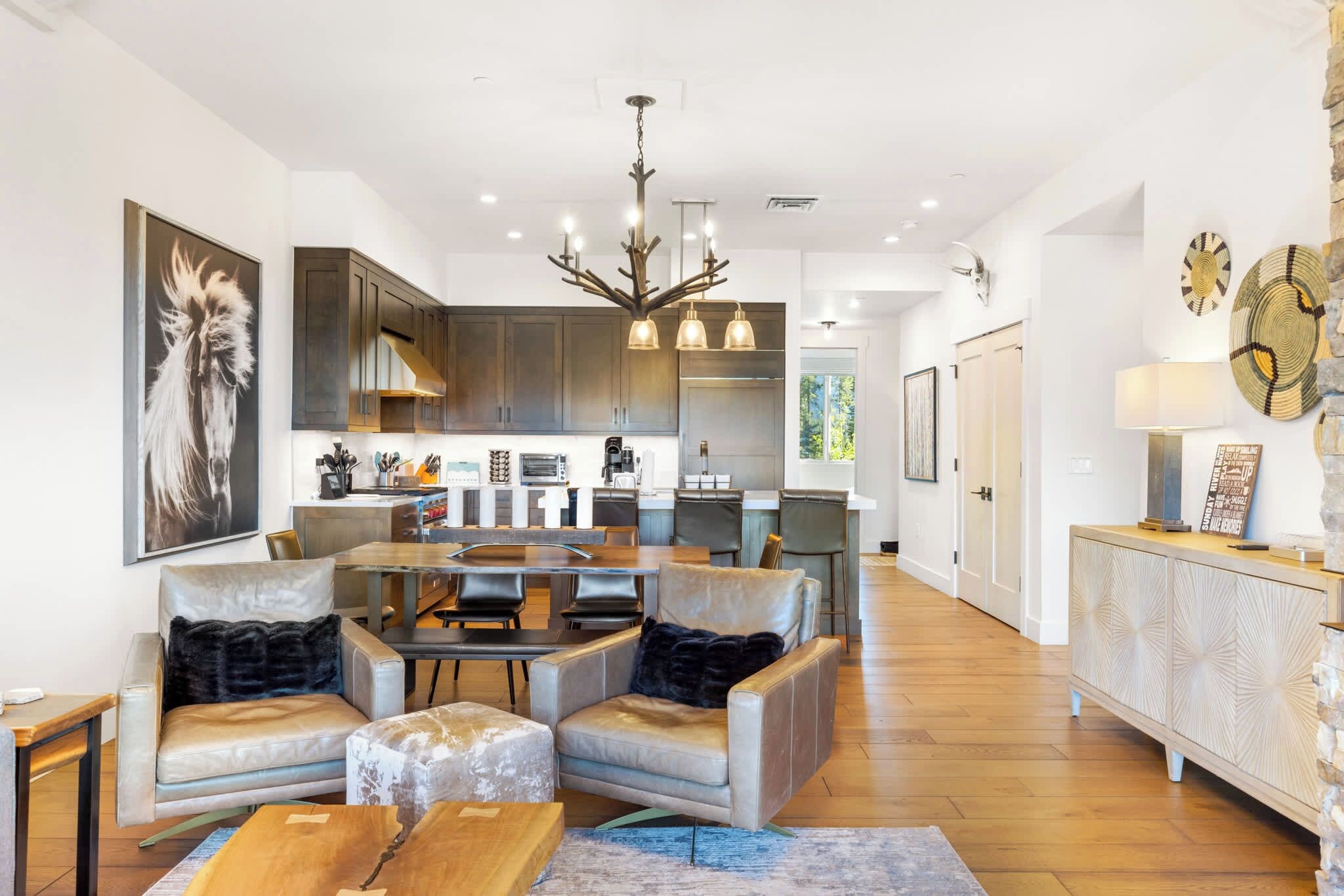 A spacious living area with leather sofas, a wooden coffee table, and a dining table with chairs. A kitchen with dark cabinets and a modern chandelier is visible in the background.