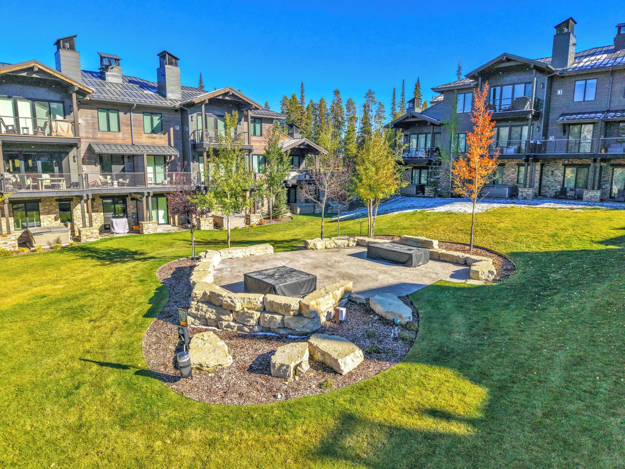 Un quartier résidentiel avec un foyer en pierre entouré de rochers et d’arbres, une pelouse bien entretenue et des maisons dotées de grandes fenêtres et de façades en pierre.