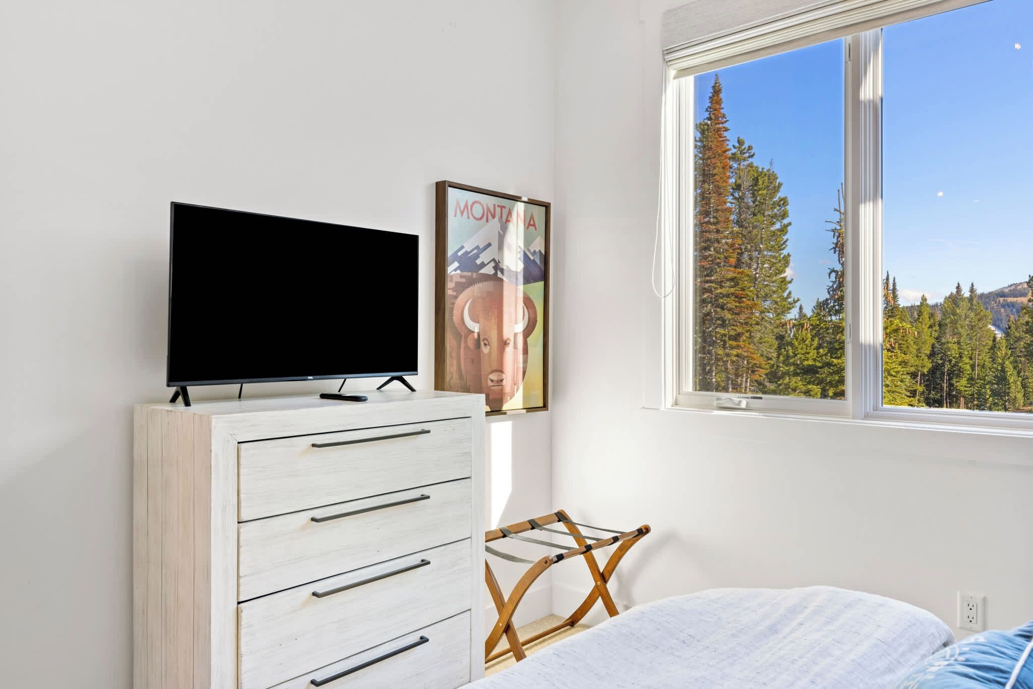 Ein Schlafzimmer mit einem Bett, einer Kommode mit Schubladen, einem Fernseher darauf, einem Poster an der Wand und einem Fenster mit Blick auf Bäume und Berge.