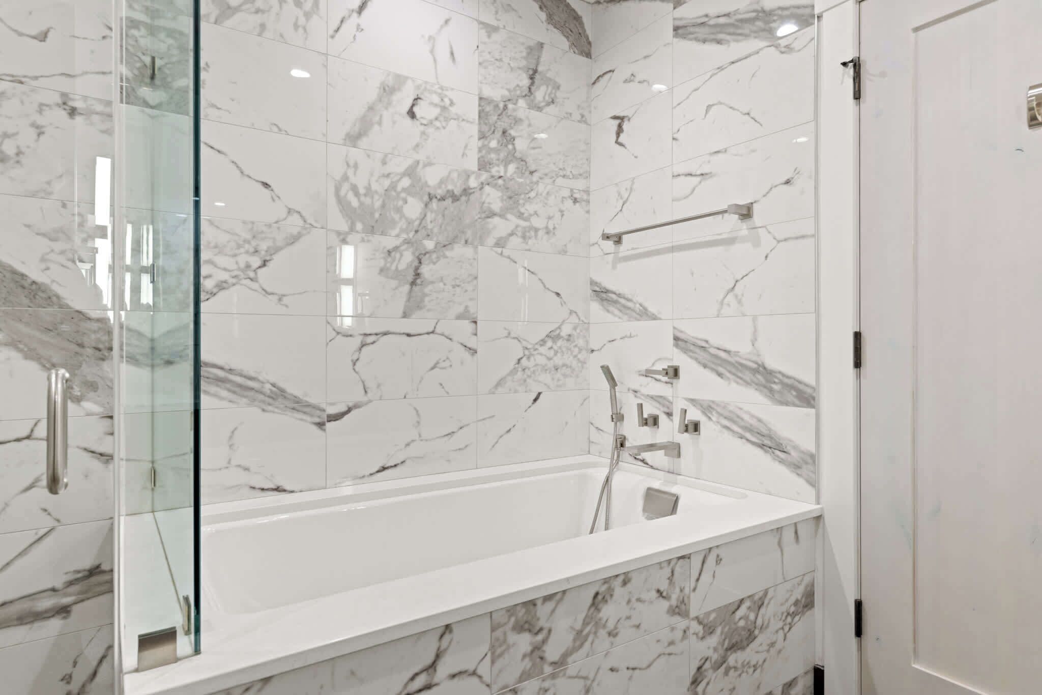 A modern bathroom with a marble-tiled shower enclosure, a glass door, and a white bathtub.