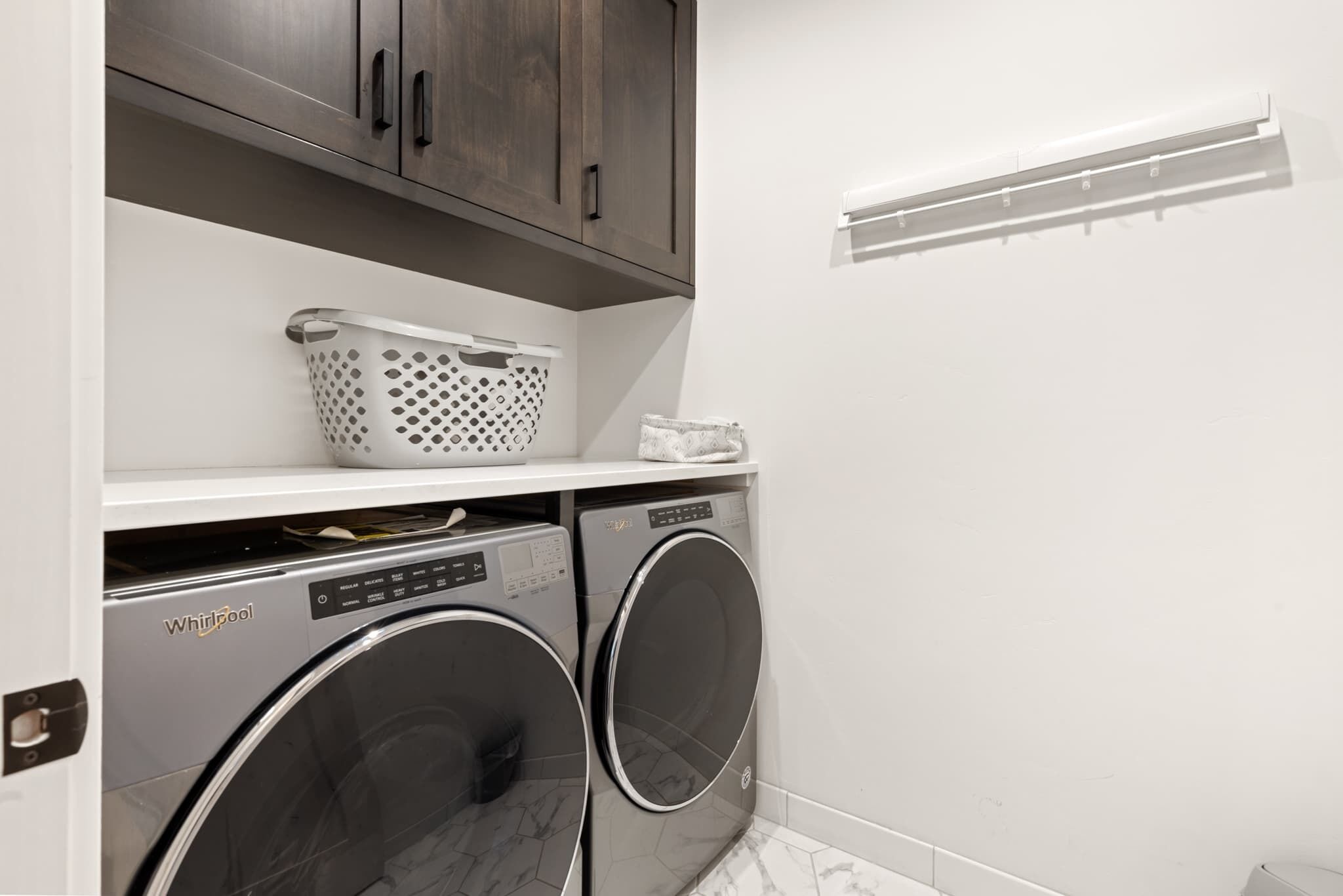 Un espace buanderie comprenant deux lave-linge Whirlpool à chargement frontal, une étagère blanche avec un panier à linge et un mur blanc doté d’une bouche d’aération.