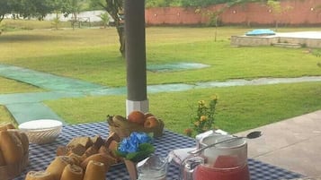 Una mesa al aire libre con mantel a cuadros, cestas de pan, frutas, una jarra de leche y un vaso de bebida roja.