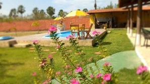 A pool area with a covered seating area, a small building, and a grassy lawn with pink flowers. - Pousada Sítio Boas Novas  (Caucaia)