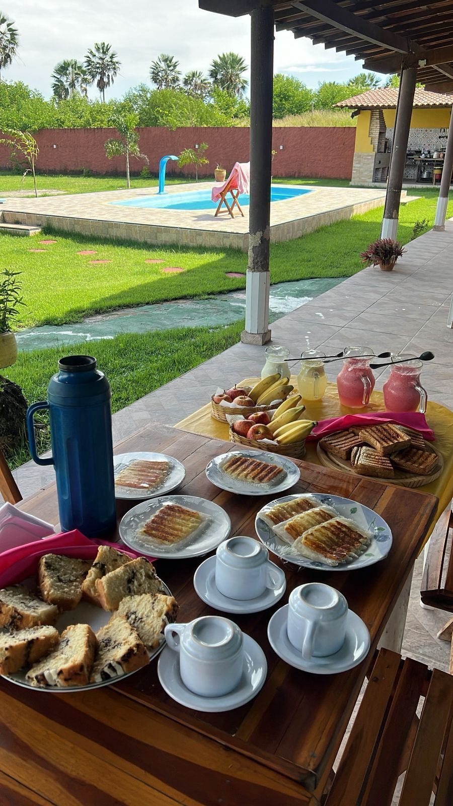 An outdoor breakfast setup with a pool view, including bread, fruits, and beverages.