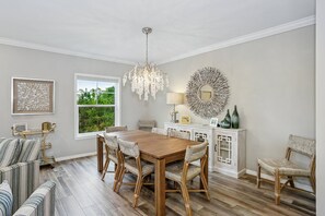 A dining area with a wooden table, wicker chairs, a chandelier, and a view of greenery through a window.