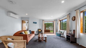 A spacious living room with a sofa, a coffee table, and a wicker chair. There is a large window with curtains, a door leading outside, and a clock on the wall. - Hideaway on Edgecumbe (Tauranga)