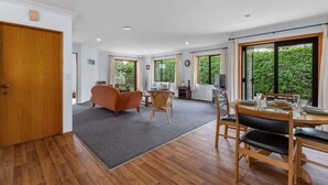 A spacious living room with a sofa, a dining table set for four, and a large window offering a view of greenery. - Hideaway on Edgecumbe (Tauranga)