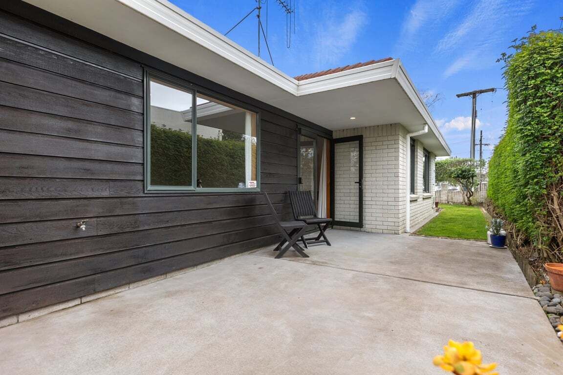 A modern house with a dark exterior, a large window, and a small patio with two chairs.