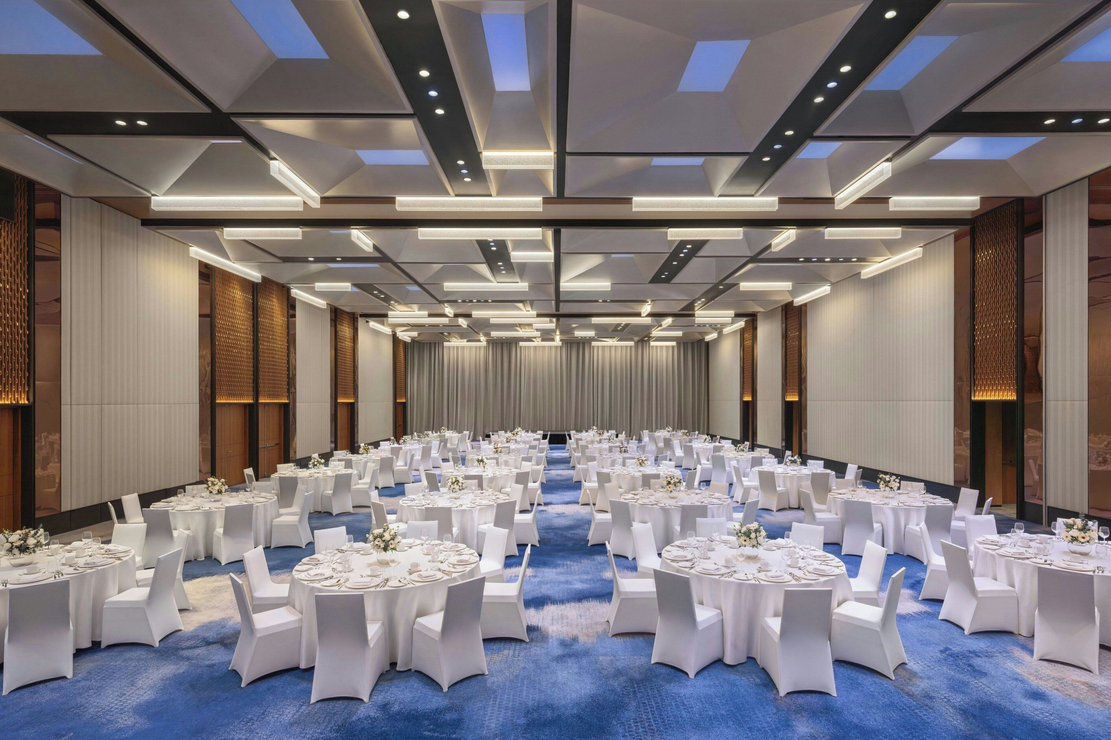 Une grande salle de conférence avec des tables rondes, des chaises blanches et un tapis bleu.