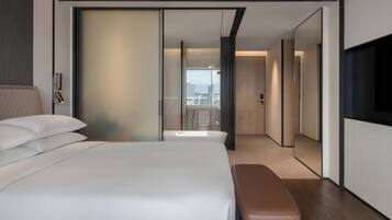 A modern hotel room with a large bed, a bench, a TV, and a view of the city.
