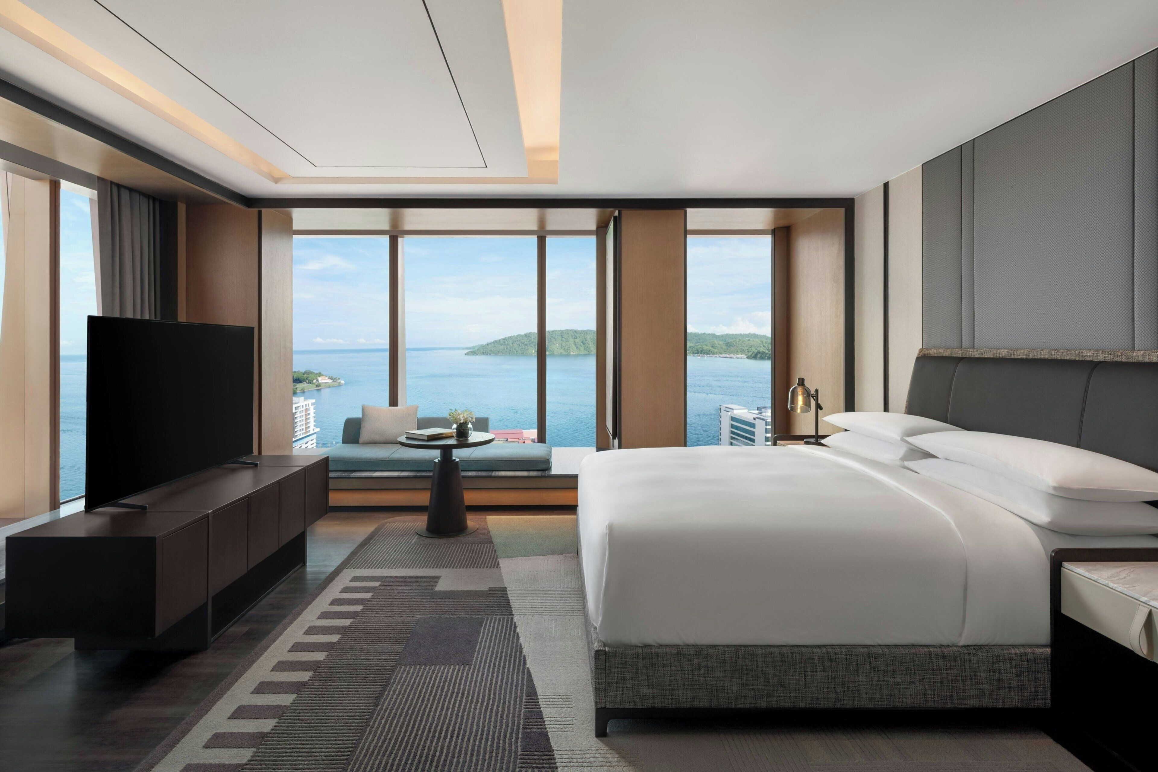 Una habitación de hotel moderna con una cama grande, un televisor de pantalla plana, un área de descanso y vistas al mar, además de un ventanal amplio.