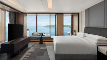 Una habitación de hotel moderna con una cama grande, un televisor de pantalla plana, un área de descanso y vistas al mar, además de un ventanal amplio.