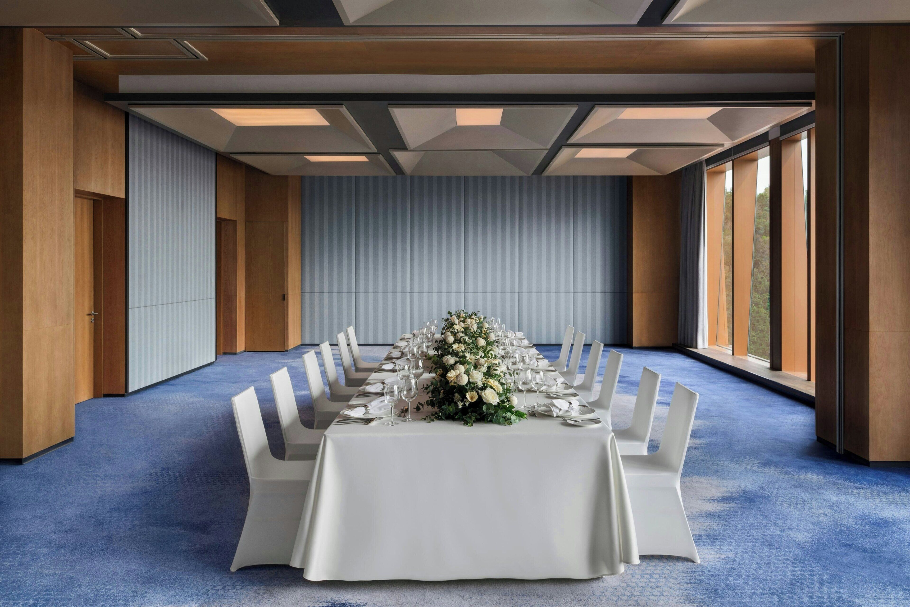 A conference room with a long table set for a formal event, flanked by tall chairs, a blue carpet, and large windows.