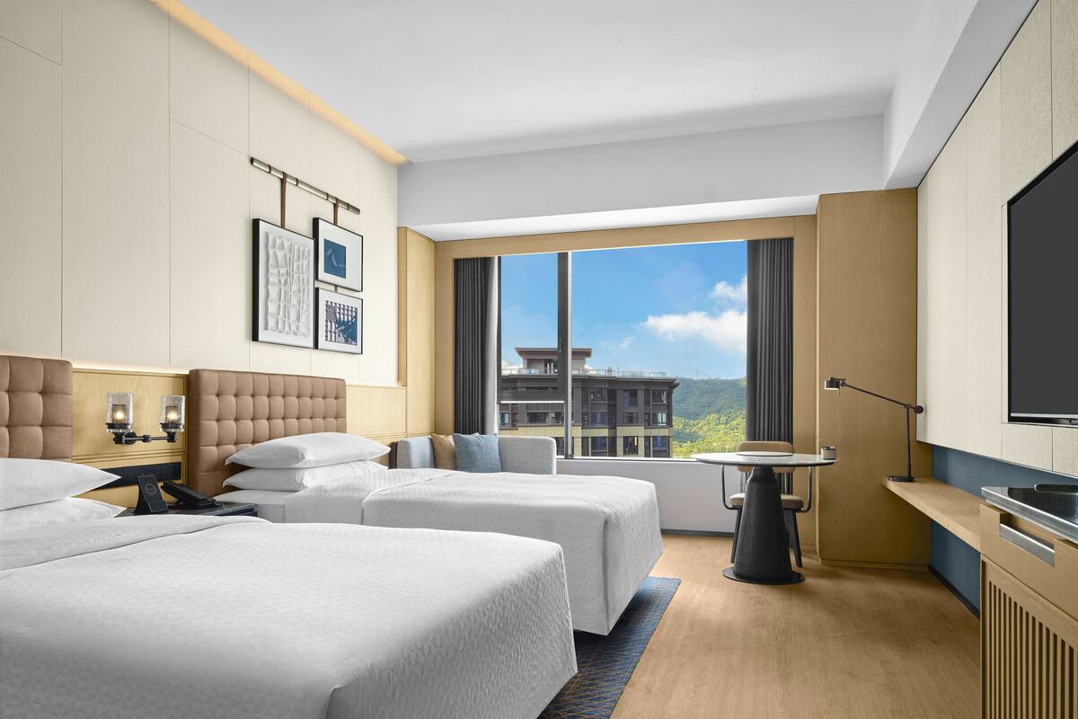 Een moderne hotelkamer met twee bedden, een groot raam met uitzicht op de stad, een kleintje tafeltje en een flatscreen televisie.