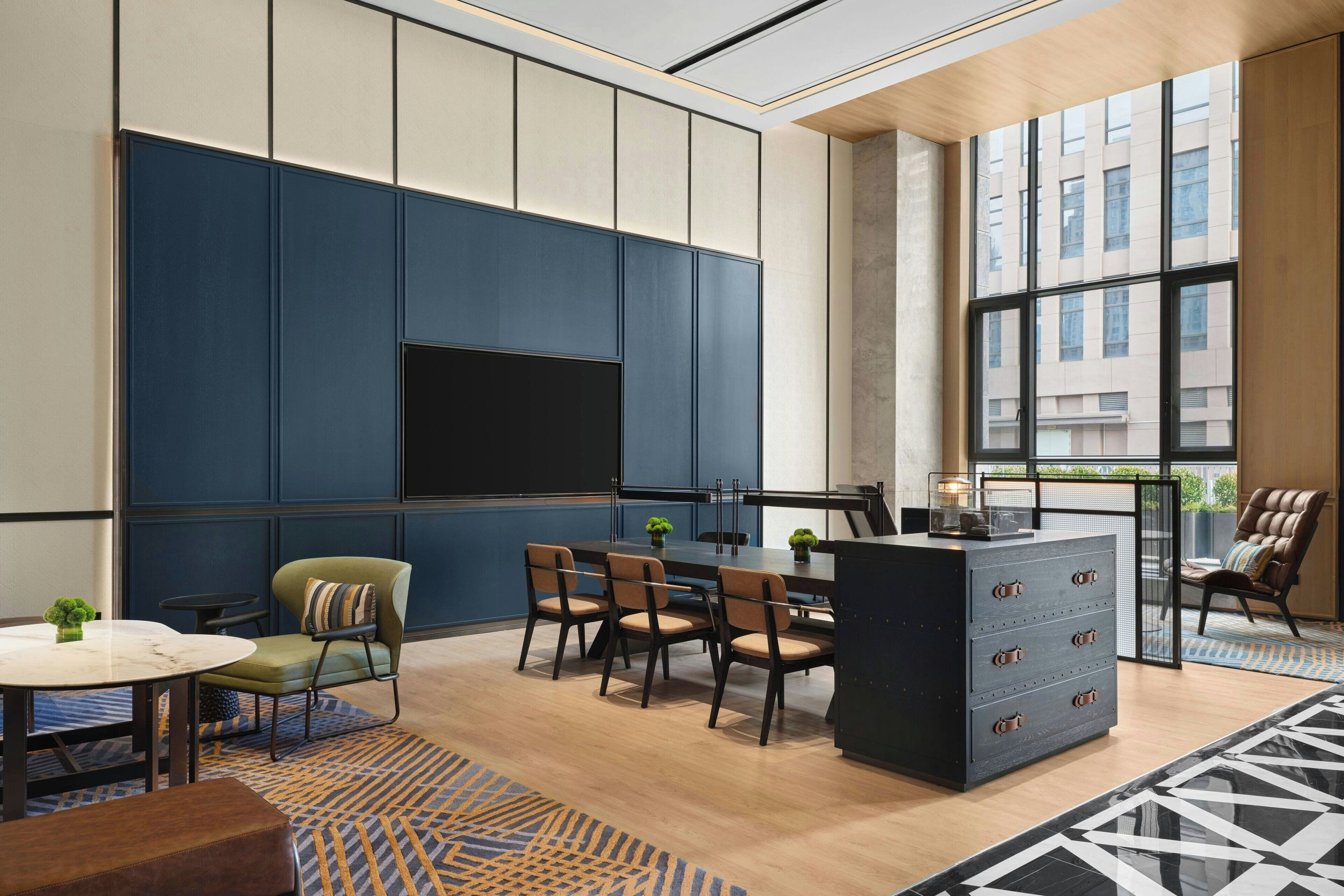 A modern hotel room with a large window, a flat-screen TV mounted on the wall, a dining area with a table and chairs, and a small seating area with a sofa and armchairs.