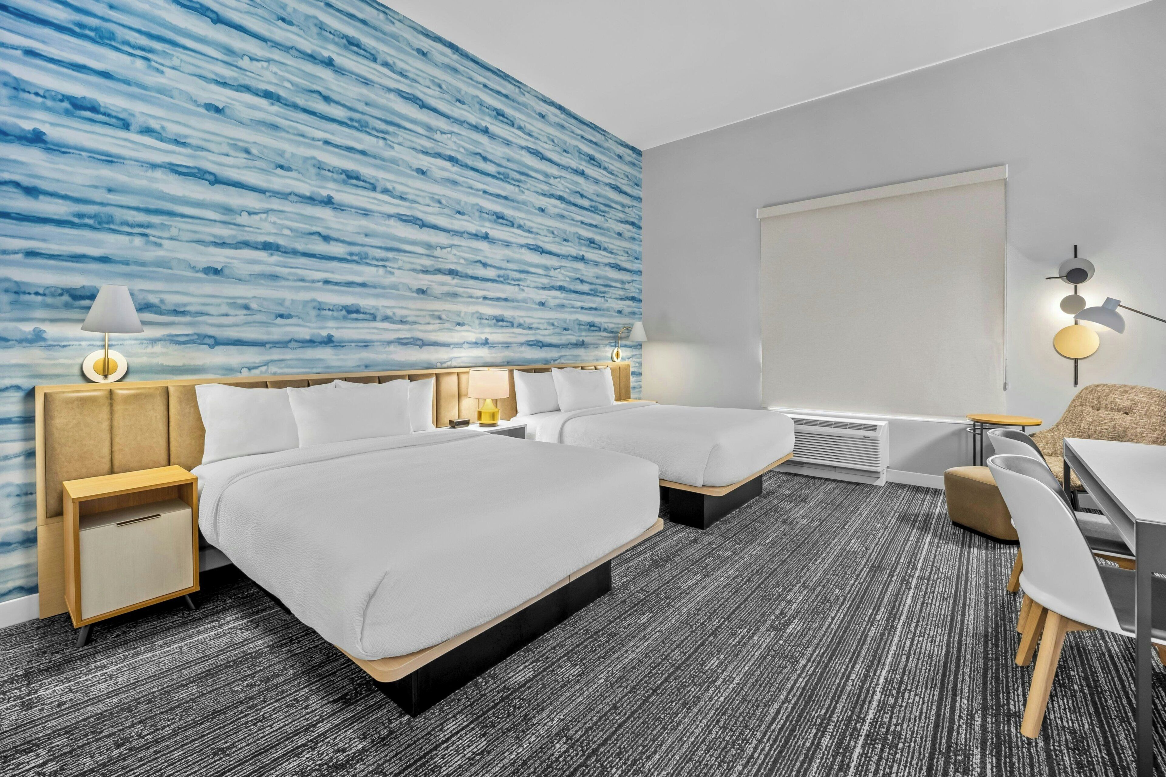 Quarto de hotel com duas camas, uma escrivaninha, uma cadeira, uma luminária na parede e uma janela grande.