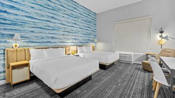 Quarto de hotel com duas camas, uma escrivaninha, uma cadeira, uma luminária na parede e uma janela grande.
