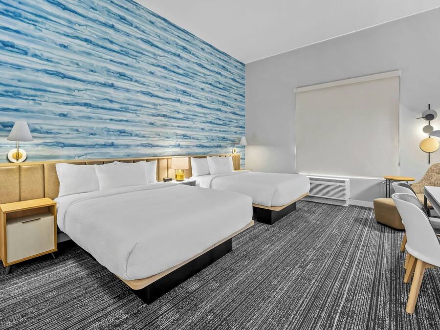 Quarto de hotel com duas camas, uma escrivaninha, uma cadeira, uma luminária na parede e uma janela grande.