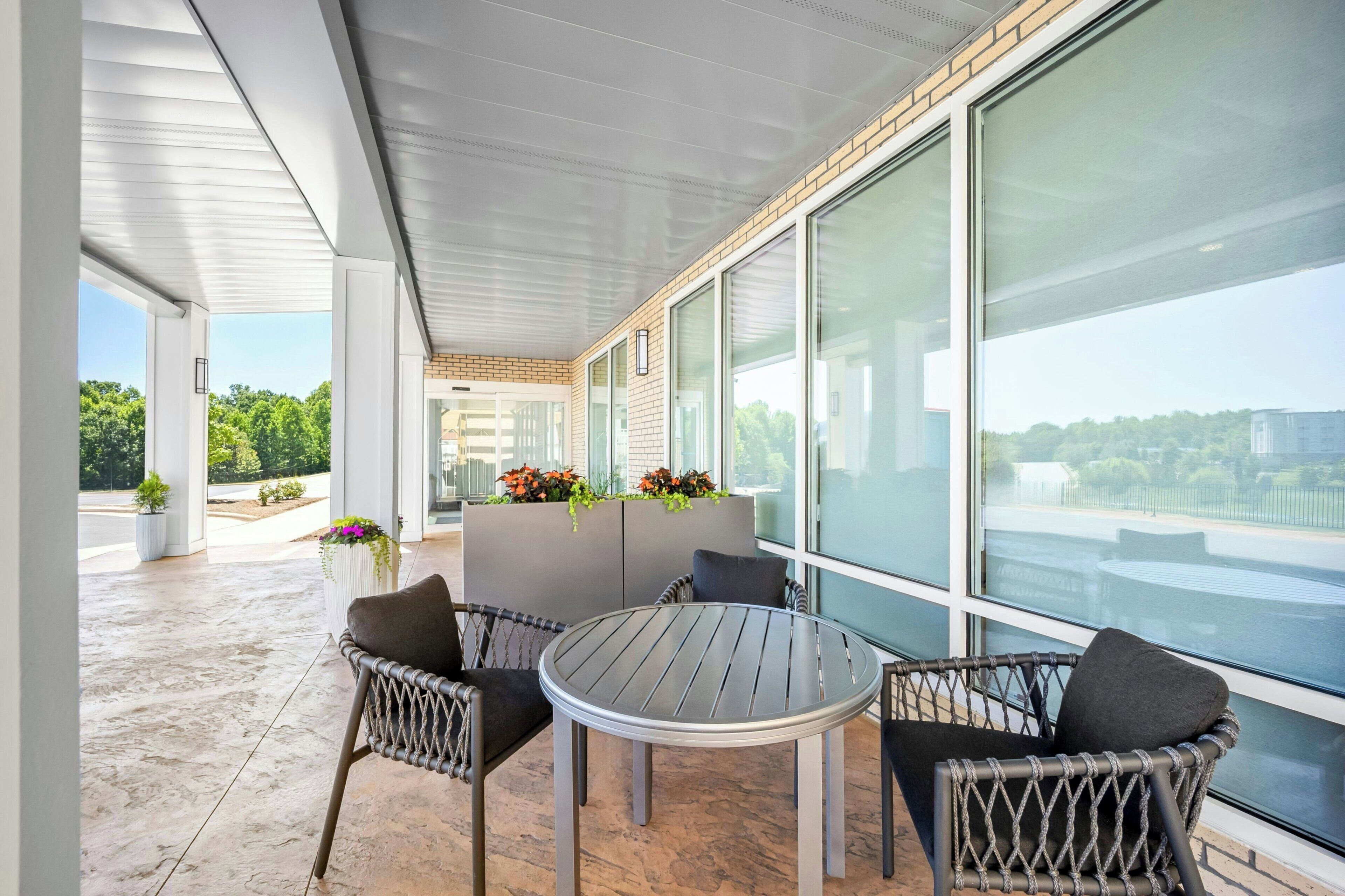 A modern outdoor seating area with a round table and wicker chairs, large windows, and potted plants.