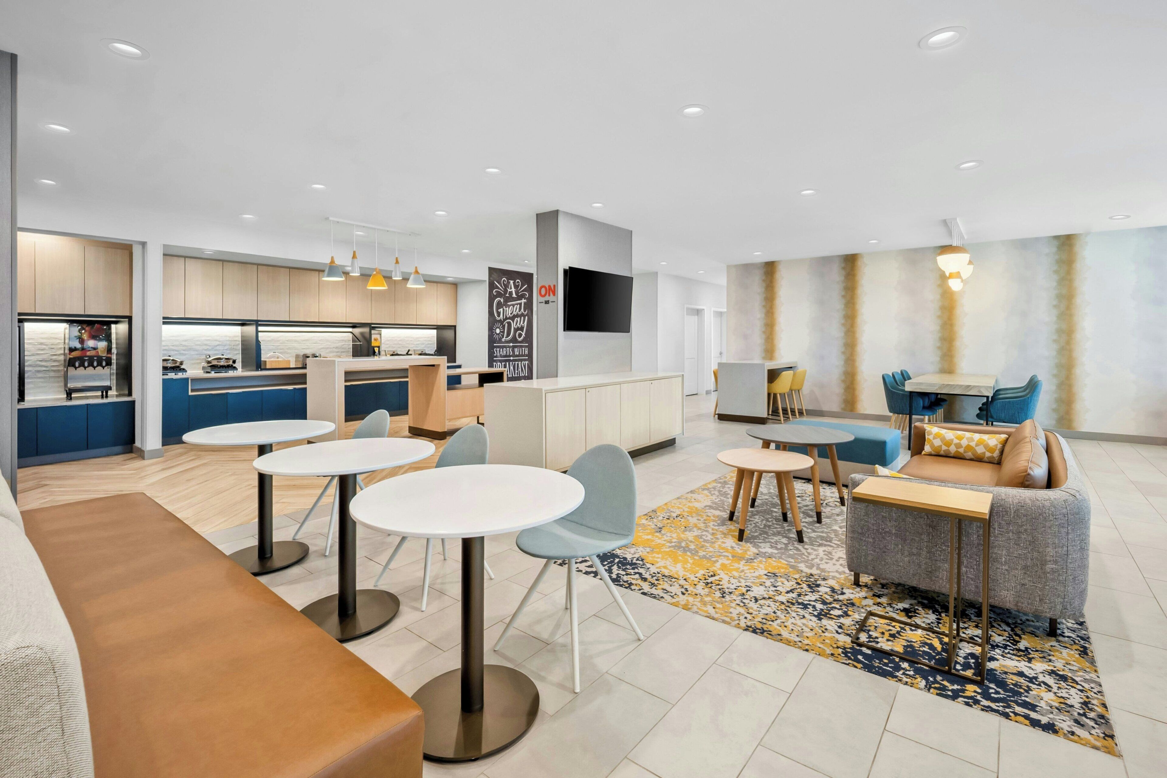 A modern hotel lobby with a kitchen area, seating arrangements, and a flat-screen TV mounted on the wall.