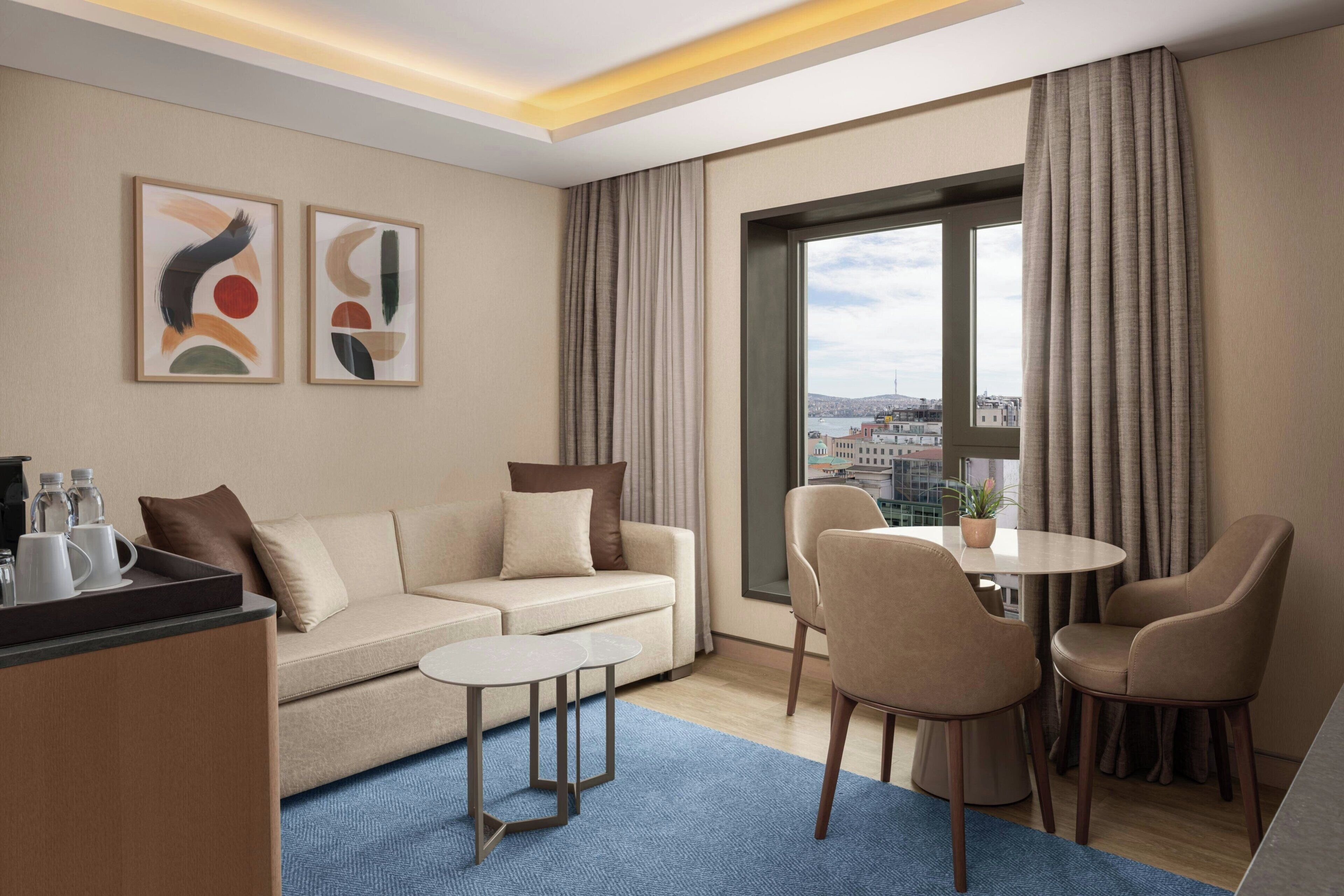 A modern hotel room with a sofa, two chairs, a small table, a coffee station, and a large window with curtains.