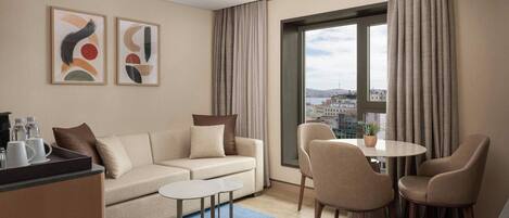 A modern hotel room with a sofa, two chairs, a small table, a coffee station, and a large window with curtains.