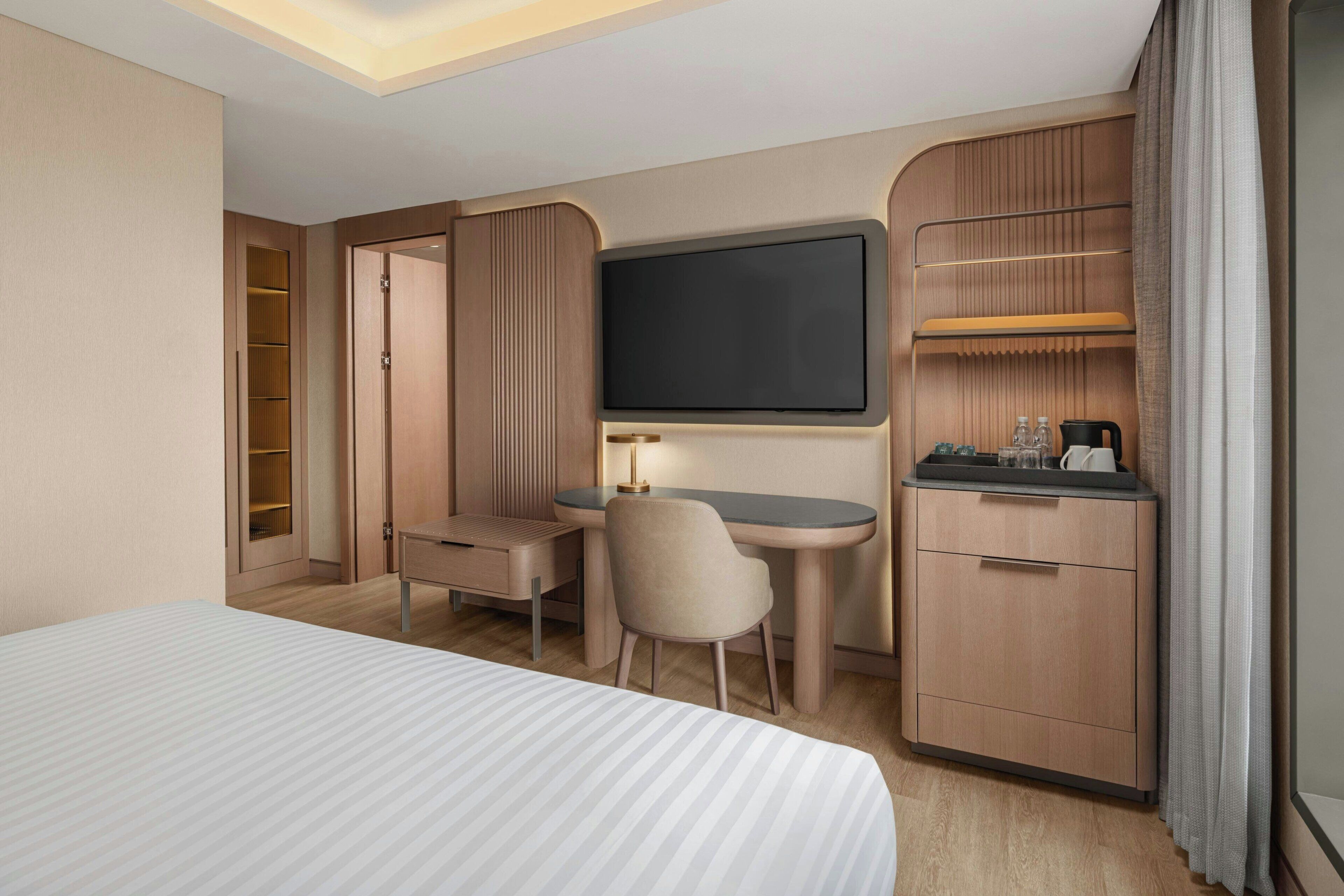 A hotel room with a bed, a desk, a chair, a TV, a minibar, and a wardrobe.